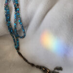 Blue glass Islamic prayer beads placed on soft fabric with a rainbow light reflection nearby.