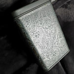Decorative silver patterned tin container resting on soft dark fabric.