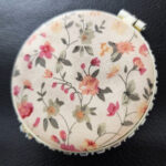 Floral compact mirror with fabric design placed on a dark textured surface.