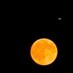 A glowing orange full moon in a dark sky with small aircraft lights visible nearby.