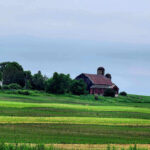Old rustic barn and silo standing on rolling green farmland under an overcast sky.