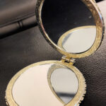 Open compact mirror showing reflection with detailed metal frame on a dark textured surface.