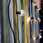 Warm outdoor string lights hanging along a wooden fence in a backyard setting.