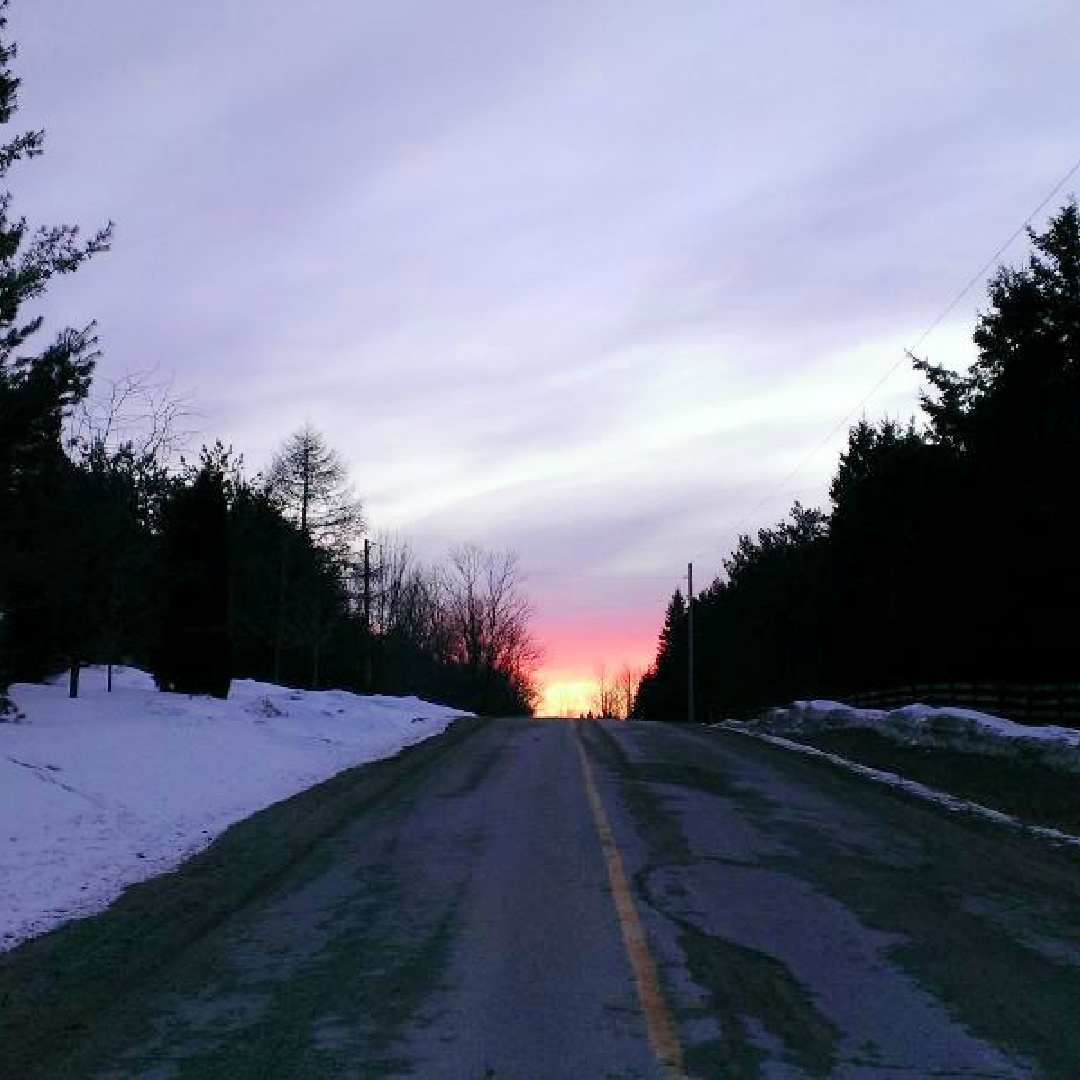 Snow-lined road leading toward a glowing sunset under a dramatic cloudy sky.