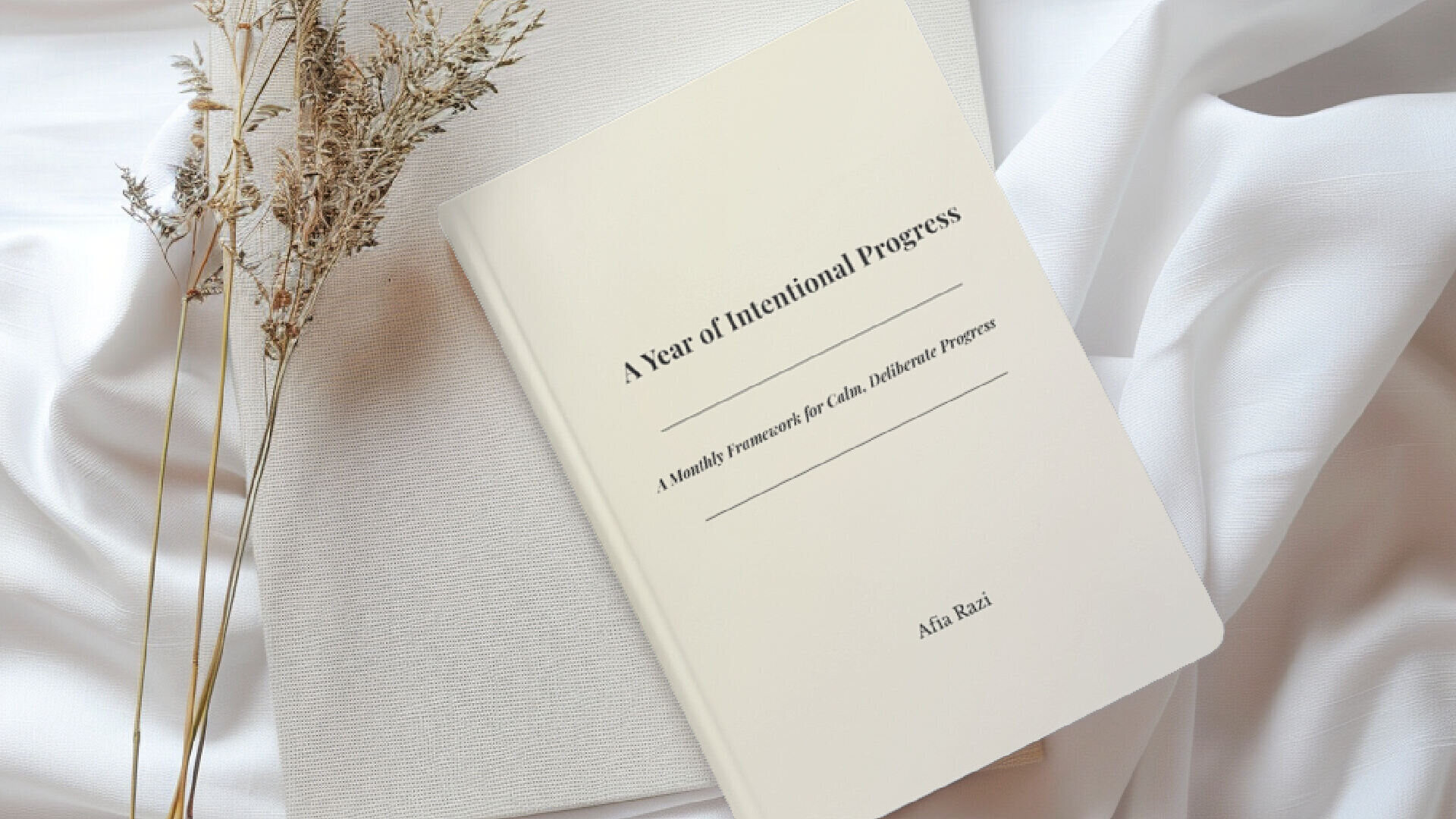 A Year of Intentional Progress book styled on soft white fabric beside a notebook and dried stems