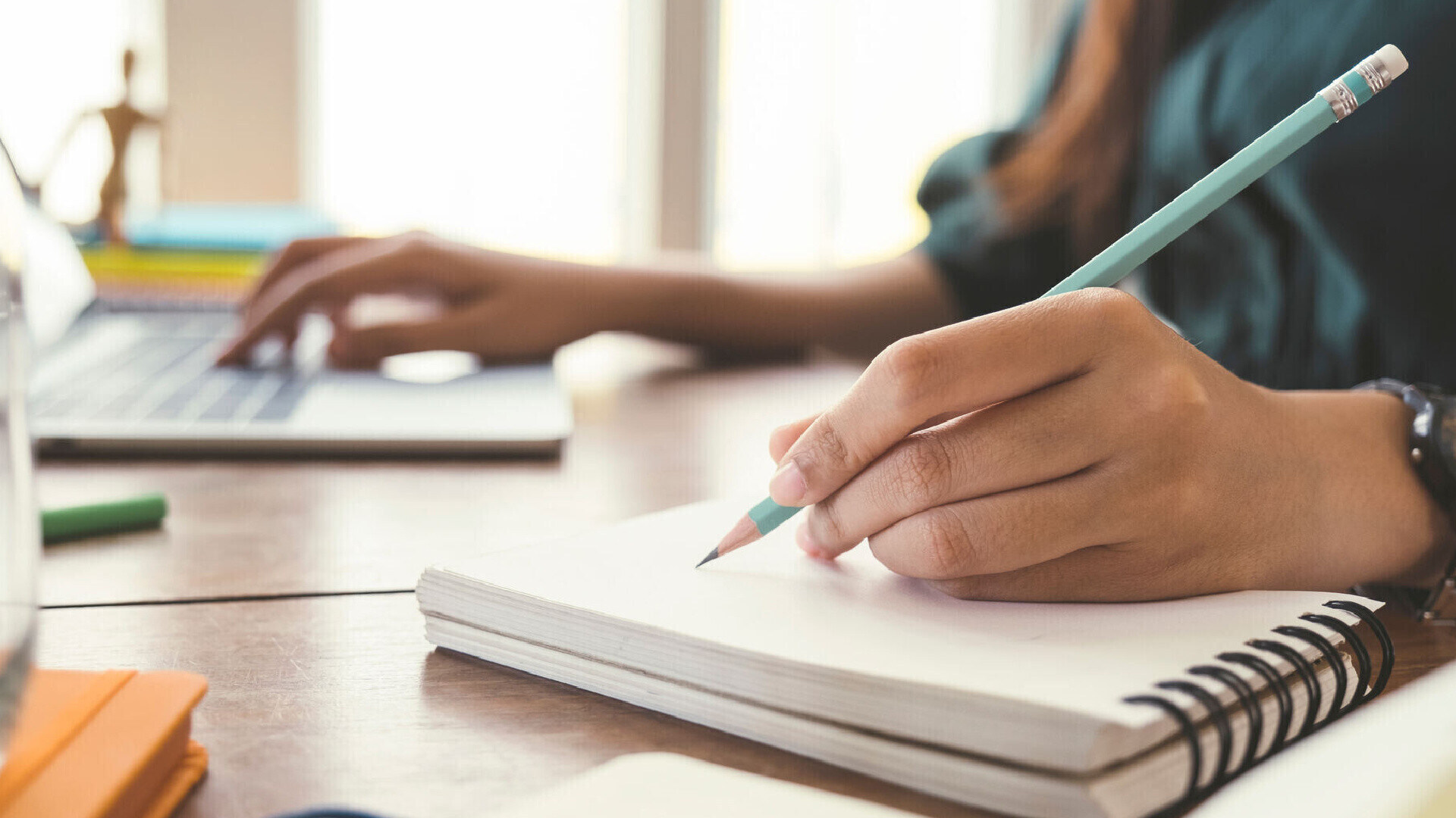 Person writing blog content in a notebook next to a laptop in a minimal workspace