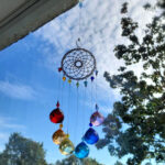 Multicoloured glass prism suncatcher hanging by a window against a blue sky with soft clouds and tree branches