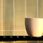 White ceramic cup resting on a metal rack against tiled wall, illuminated by warm sunlight with soft shadow