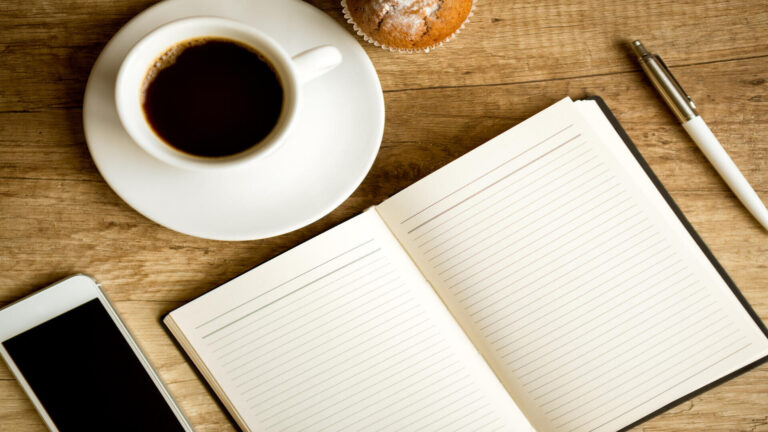 Open notebook beside a cup of coffee, smartphone, and pen on a wooden desk, representing business planning and sustainable growth.