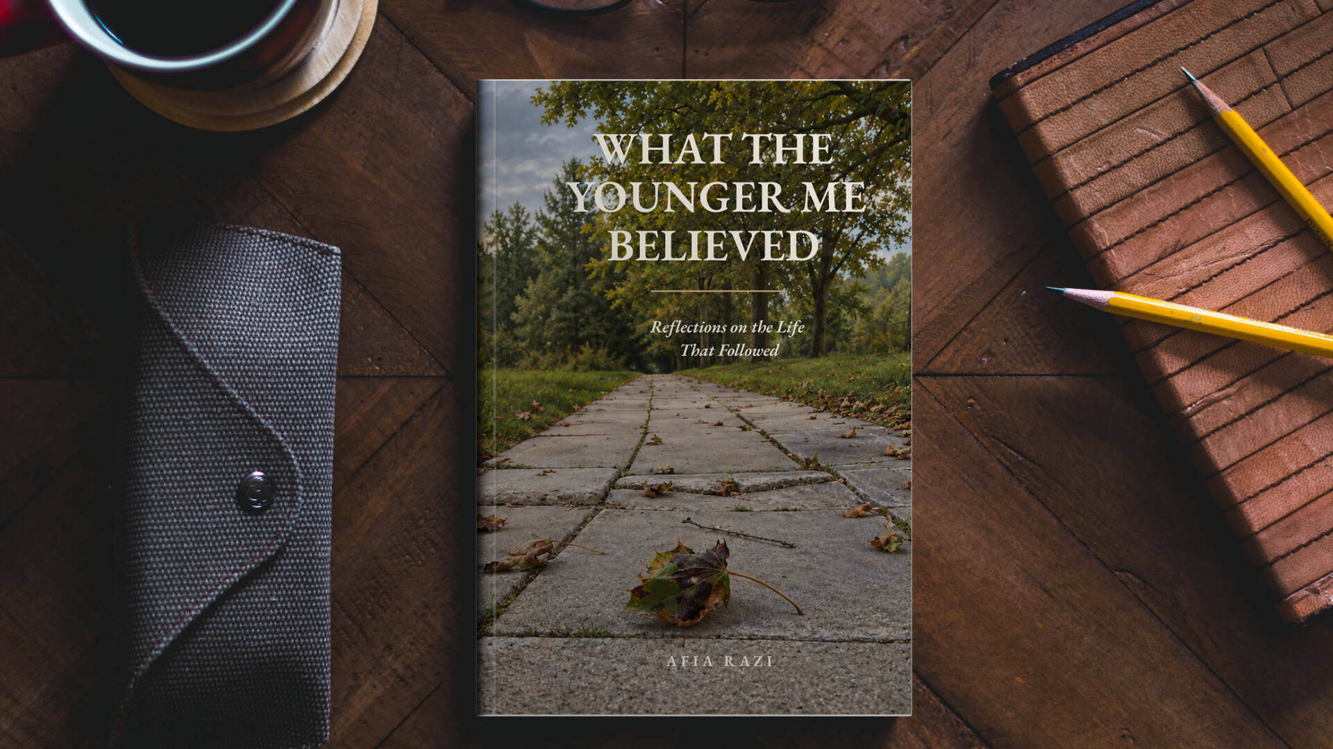 Mockup of Afia Razi’s book What the Younger Me Believed on a wooden desk beside a notebook, pencils and a cup.